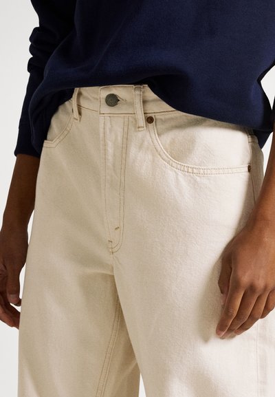 Ανοιχτό μπεζ τζιν με ψηλή μέση, με εμφανείς ραφές, πέντε τσέπες και μεταλλικό κουμπί. Συνδυάζεται με μπλε πουλόβερ του ναυτικού.