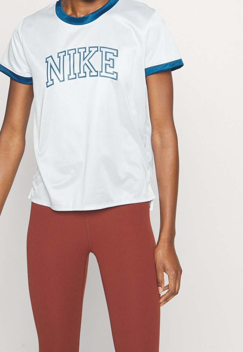 Kvinna klädd i en vit Nike-t-shirt med blåa detaljer och bruna leggings, stående mot en helt vit bakgrund.