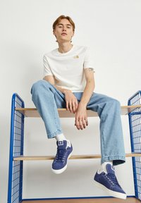T-shirt branca de manga curta com um logótipo dourado, jeans azul claro e ténis azul-marinho com detalhes brancos, a sentar-se numa prateleira de madeira dentro de uma estrutura de arame azul.