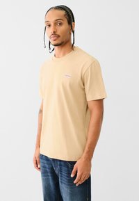 Beige pamut póló rövid ujjal, kerek nyakkivágással és egy kis piros logóval a bal mellrészen. Kék farmerrel viselve.