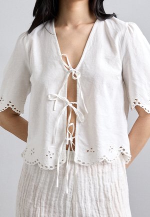 Femme portant un chemisier blanc à manches courtes avec des bords festonnés à œillets et trois liens à l'avant sur un pantalon texturé clair.