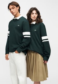 Deux mannequins portent des polos vert foncé à manches longues avec des cols blancs et des rayures sur les manches ; l'une porte un pantalon blanc, l'autre une jupe plissée beige.