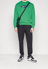 Felpa verde con logo, pantaloni neri e scarpe da ginnastica in bianco e nero. L'outfit include una piccola borsa a tracolla nera.