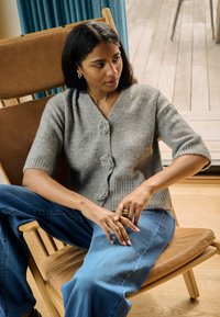 Cardigan gri cu mâneci scurte, cu nasturi în formă de floare și tiv răsfrânt, asortat cu blugi albaștri din denim, purtat în timp ce stă pe un scaun de lemn.