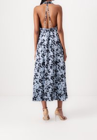 Robe longue à col licou, présentant un imprimé abstrait bleu et noir, un dos nu et une silhouette fluide. Talons transparents complètent le look.