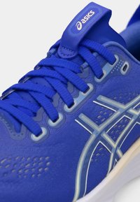 Zapatilla deportiva azul con una parte superior texturizada, detalles de malla y acentos blancos en contraste. Presenta cordones planos y una etiqueta en la lengüeta con la marca.