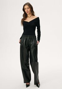 Kobieta w czarnej bluzce odkrywającej ramiona, luźnych czarnych skórzanych spodniach, czarnych szpilkach z zaostrzonym noskiem i złotym naszyjnikiem na jednolitym jasnym tle.