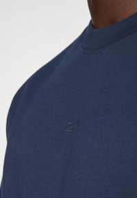 Marineblå hettegenser med klassisk passform, ribbestrikket halslinning og diskret brodert logo på brystet, laget av myk, strukturert stoff.