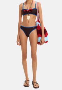 Brązowy bikini z niebieskimi akcentami, z topem bandeau i wysokim stanem. Modelka nosi czerwone okulary przeciwsłoneczne i trzyma torebkę z frędzlami w wielu kolorach.