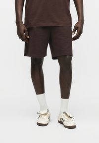 Άνδρας που φοράει σκούρα καφέ σορτς, λευκές κάλτσες μέχρι τον αστράγαλο και λευκά αθλητικά παπούτσια με μαύρες ρίγες, στέκεται μπροστά σε ουδέτερο φόντο.