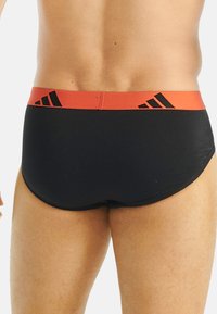 Czarne spodenki kąpielowe z czerwonym elastycznym pasem w talii ozdobionym czarnymi paskami i logo. Gładka struktura, dopasowany krój i minimalne przykrycie.