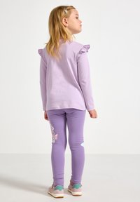 Lyslilla langærmet skjorte med flæsekantede skuldre, kombineret med lyslilla leggings med sommerfugleprint og lyserøde sko med grønne detaljer.