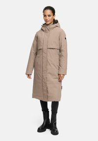Longue doudoune beige avec capuche, fermeture éclair à l'avant et poches à rabat. Portée sur un pantalon noir et des bottes noires robustes. Texture lisse et isolée.