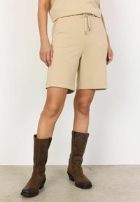 Shorts beige in misto cotone con vita elastica e cordino, abbinati a stivali in pelle marrone al di sopra del ginocchio con tacco spesso.