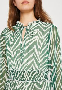 Grön och vit mönstrad blus med zebraranddesign, som har en volangkrage, knytning och smockad midjedetalj.