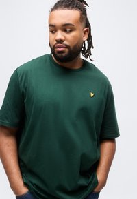 Άνδρας με ράστα και μούσι, που φοράει σκουροπράσινο μπλουζάκι με κίτρινο λογότυπο πουλιού, με τα χέρια στις τσέπες, κοιτάζει αριστερά μπροστά σε απλό φόντο.