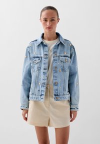 Giacca di jeans azzurro chiaro con borchie oro e argento, chiusura con bottoni, due tasche frontali e una vestibilità leggermente rilassata. Abbinata a una t-shirt leggera.