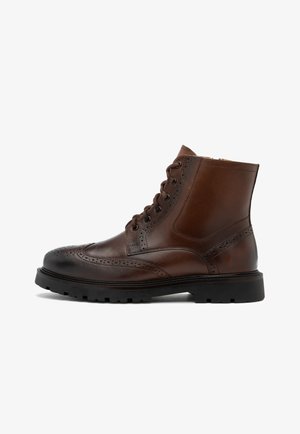 Brązowe skórzane botki na kostkę z detalami w stylu brogue, okrągłym noskiem, sznurowanym zapięciem i grubą czarną gumową podeszwą. Gładka faktura z kontrastowymi akcentami.