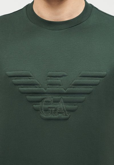 Σκούρο πράσινο μπλουζάκι με στρογγυλή λαιμόκοψη, με κεντημένο λογότυπο αετού και τα γράμματα "GA" στο κέντρο του στήθους.