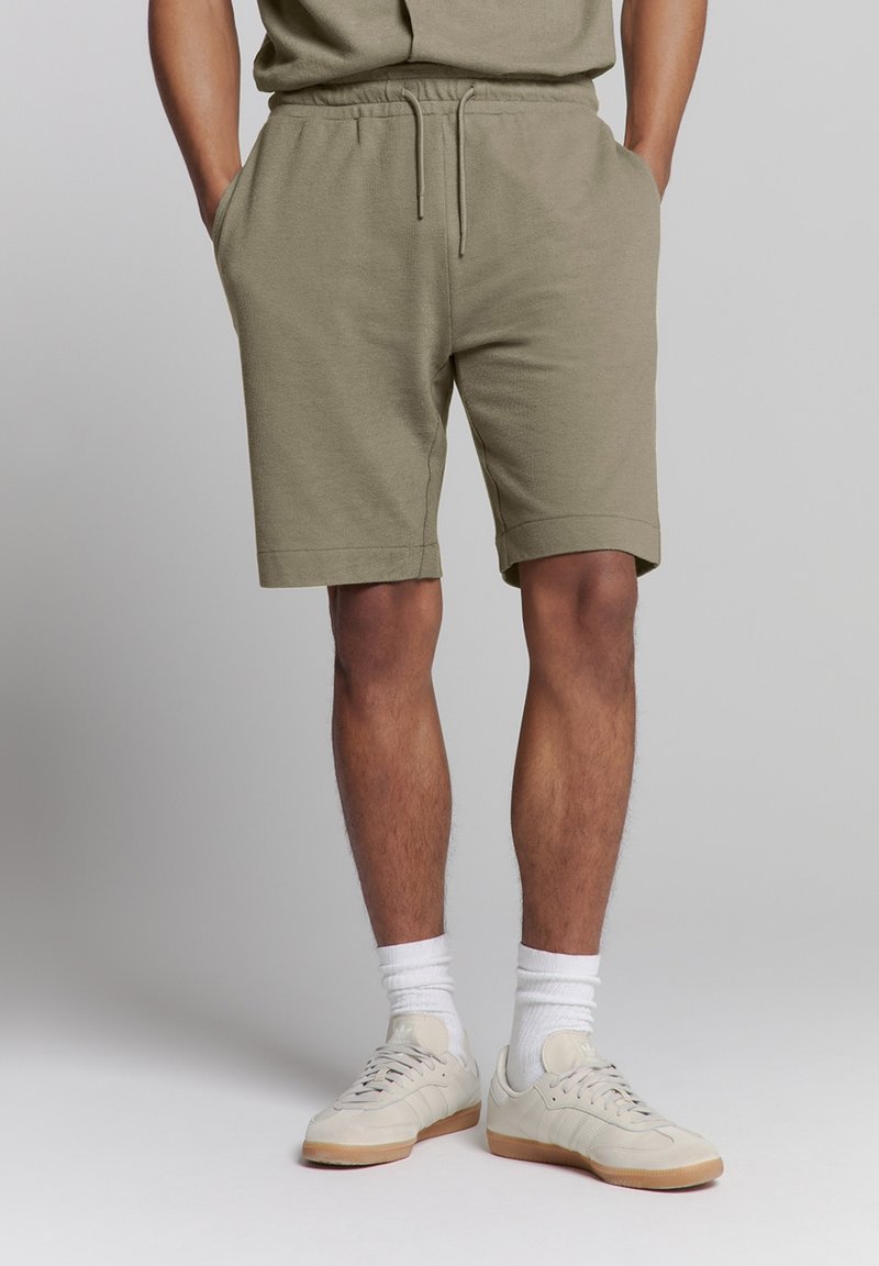 Khaki sweatshorts met een elastische tailleband en een trekkoord, voorzien van zijzakken en een opgerolde zoom. Gecombineerd met lichtgekleurde sneakers.