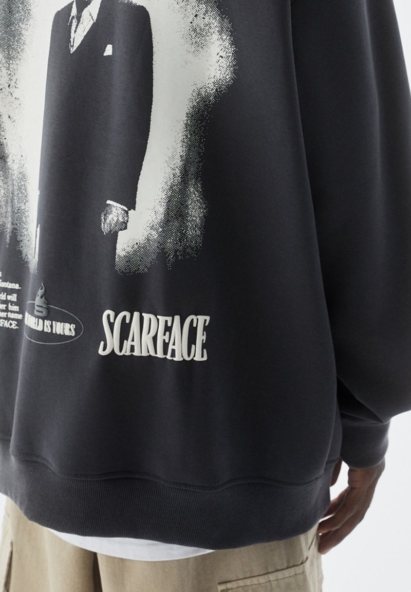 Sweat-shirt noir avec graphisme en noir et blanc d'un homme en costume et le texte "Scarface" et "The World Is Yours" dans le dos.