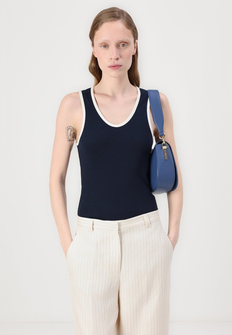 Granatowa bezrękawnik z białym wykończeniem, o żeberkowej fakturze. W połączeniu z jasnymi, w paski szerokimi spodniami i niebieską torebką. Gładkie białe tło.