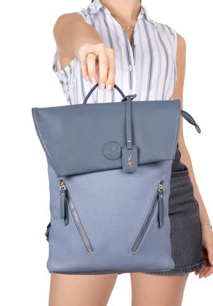 Femme tenant un sac à dos bleu avec rabat en cuir et deux poches verticales zippées à l'avant, portant un débardeur rayé et une jupe noire.