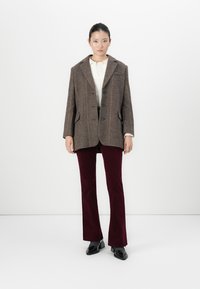Vrouw staande in een bruine geruite blazer, witte top, bordeauxrode broek en zwarte vierkante schoenen tegen een effen witte achtergrond.
