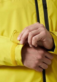 Giacca impermeabile gialla con polsini regolabili in velcro. Inquadratura ravvicinata di mani che chiudono il polsino, mostrando la trama liscia del tessuto e l'hardware a contrasto.