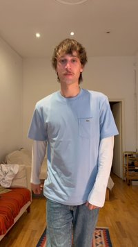 Νεαρός άνδρας με σγουρά μαλλιά, ντυμένος με στρωματωτά ανοιχτό μπλε και λευκά πουκάμισα και τζιν, στέκεται σε ένα σαλόνι με ξύλινο πάτωμα και καναπέ.