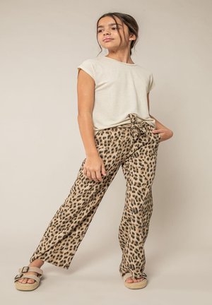 Jong meisje in een beige shirt met korte mouwen, panterprint broek met strikceintuur en beige sandalen, staand met één hand in de zak.