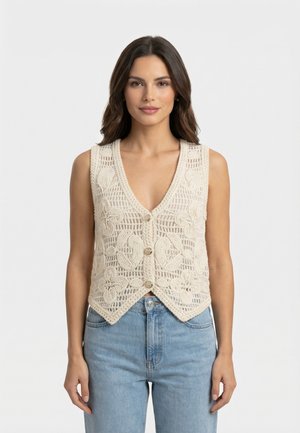 Kobieta w kremowej, szydełkowej kamizelce z kwiatowymi wzorami i guzikami, w połączeniu z wysokim stanem jasnoniebieskich jeansów, stojąca na jednolitym tle.