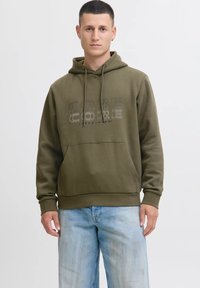Jonge man in een groene hoodie met de tekst "CORE" en lichtblauwe jeans, staande tegen een effen lichte achtergrond, met het gezicht naar voren.