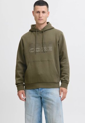 Giovane uomo che indossa un hoodie verde con la scritta "CORE" e jeans blu chiaro, in piedi contro uno sfondo chiaro e uniforme, rivolto in avanti.