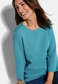 Frau mit braunem Haar, die lächelt und einen strukturierten, türkisfarbenen Pullover mit dreiviertel Ärmeln trägt, sitzt an einer weißen Wand.