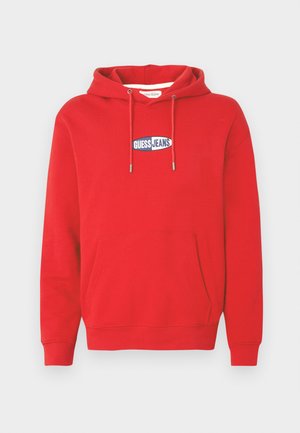 Roter Kapuzenpullover aus weichem Stoff, mit einer Fronttasche, Kapuze mit Kordelzug und einem blauen und weißen "GUESS JEANS" Logoaufnäher auf der Brust.