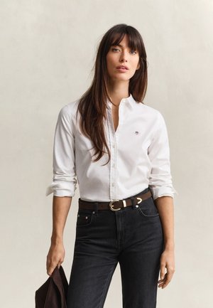 Femme aux longs cheveux bruns portant une chemise blanche boutonnée, un jean noir et une ceinture marron, tenant une veste marron foncé dans une main.
