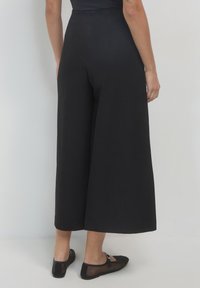 Eine Frau trägt schwarze, weit geschnittene, gestutzte Hosen und schwarze, durchsichtige Mary Jane-Schuhe. Sie steht vor einem schlichten weißen Hintergrund.