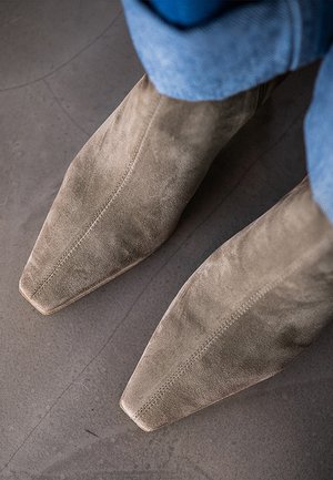 Paar taupefarbene Wildlederstiefel mit quadratischer Spitze, getragen zu blauen Jeans, von oben auf einem grauen Boden fotografiert.