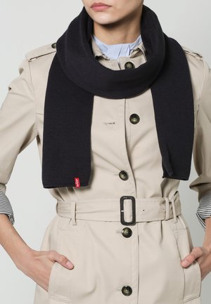 Sciarpa nera a maglia con una trama liscia, avvolta attorno al collo. Accentuata da un piccolo etichetta rossa, abbinata a un trench color beige.
