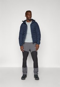 Doudoune marine avec texture matelassée, t-shirt gris clair et pantalon gris avec des sections plus foncées. Des chaussures noires complètent l'ensemble.