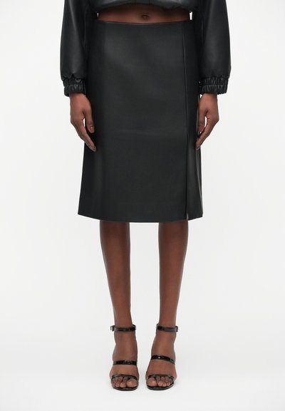Jupe midi en cuir noir avec une coupe droite, fente latérale et texture lisse. Portée avec des talons noirs à lanières.