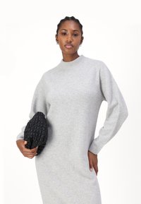 Robe tricotée grise avec un col montant et de longues manches amples, complétée par une pochette noire texturée tenue dans la main droite du modèle.
