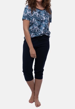 Vrouw met krullend haar die een blauw bloemig shirt met korte mouwen en zwarte cropped broek draagt, staat op blote voeten tegen een witte achtergrond.