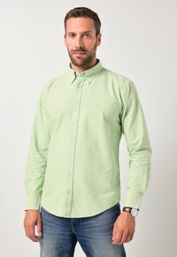 Mann, der ein hellgrünes Hemd mit Knöpfen und blaue Jeans trägt, steht vor einem schlichten hellen Hintergrund, mit einer Armbanduhr am linken Handgelenk.