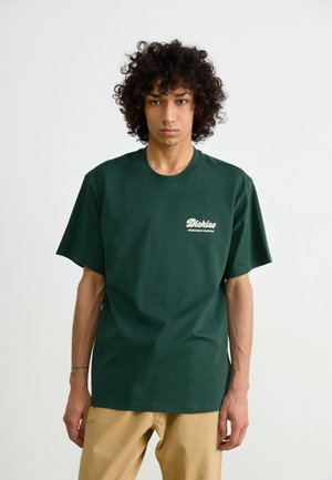 Dark green cotton t-shirt with short sleeves, featuring a white logo on the left chest. Straight fit and smooth texture. Sand-colored pants.