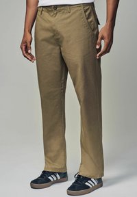Pantaloni dritti color khaki realizzati in misto cotone, con tasche laterali e chiusura a bottoni, abbinati a sneaker nere con strisce bianche.
