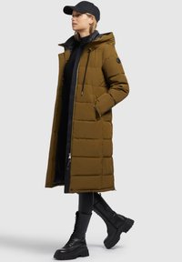 Olive green long puffer coat with a hood, featuring boxy quilting, black accents, and a front zipper. Paired with black combat boots.