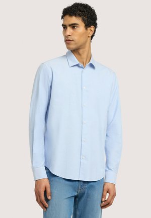 Man draagt een lichtblauw overhemd met knopen en blauwe spijkerbroek, staat met ontspannen handen langs zijn zij, kijkt naar links.