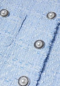 Tissu bleu clair texturé avec un motif tissé, présentant des boutons semblables à des perles entourés d'accents argentés et des bords effilochés.